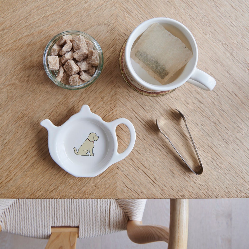 White teapot-shaped Apricot Cockapoo / Labradoodle teabag dish with a pale apricot dog illustration, beside a mug, sugar cubes and sugar tongs