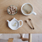 White teapot-shaped Apricot Cockapoo / Labradoodle teabag dish with a pale apricot dog illustration, beside a mug, sugar cubes and sugar tongs