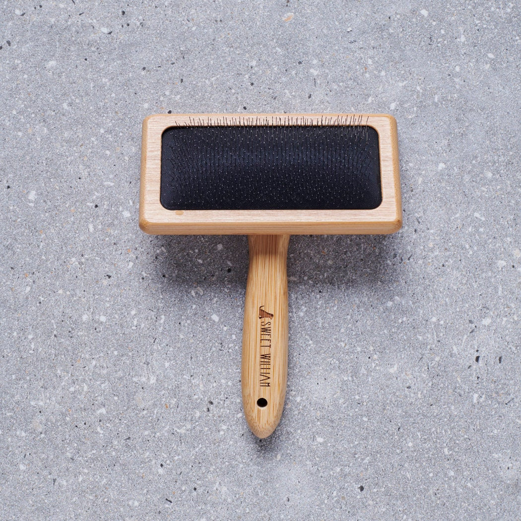 Bamboo slicker brush laid flat on grey stone surface with Sweet William branding — dog grooming gift
