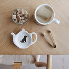 White teapot-shaped Chocolate Cocker Spaniel teabag dish with a brown dog illustration and red collar, beside a mug, sugar cubes and sugar tongs