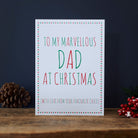White Dad Christmas card with red and green lettering reading “To My Marvellous Dad at Christmas” and “With love from your favourite child”, a star border, pine cone and red berries beside it.