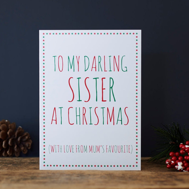 White Sister Christmas card with red and green lettering reading “To My Darling Sister at Christmas” and “With love from Mum’s favourite”, a star border, pine cone and red berries beside it.