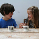 Two children sitting together at a table with ceramic “Big Brother” and “Little Sister” mugs in front of them, showing a lovely matching sibling gift